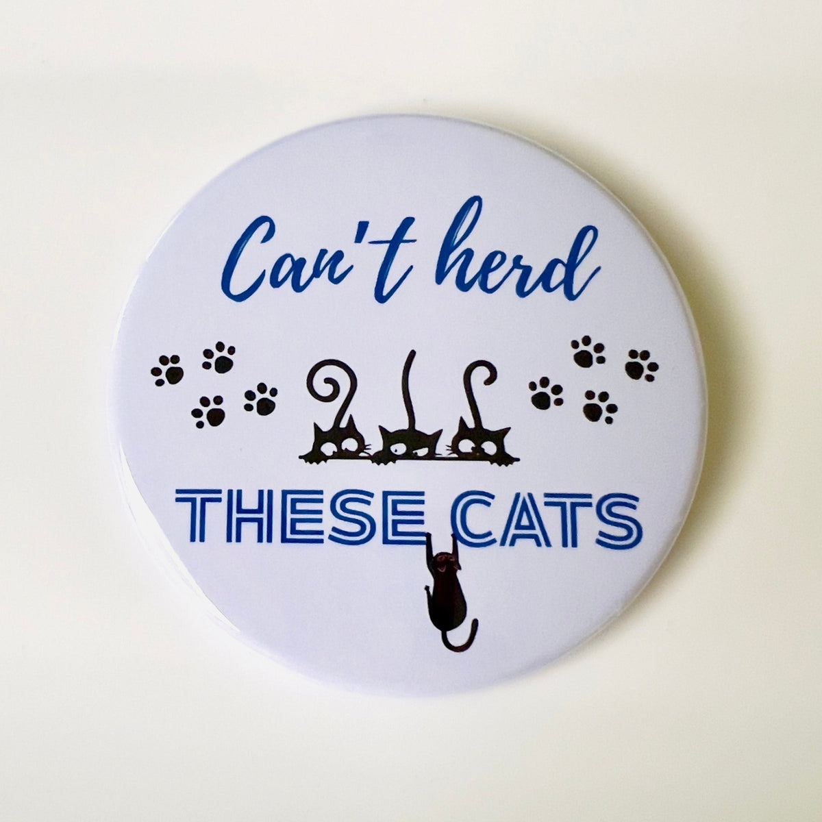 Button badge with text 'Can't herd these cats' and cat illustrations on a white background