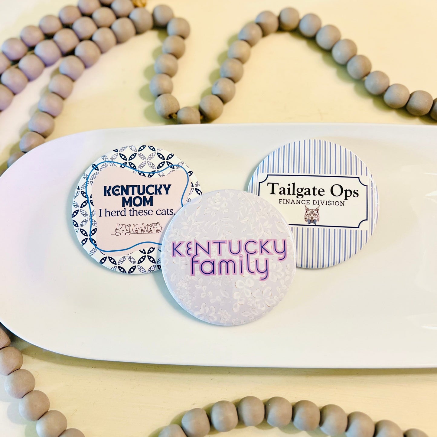 Three round Kentucky buttons with text on a white tray, surrounded by gray beaded garland