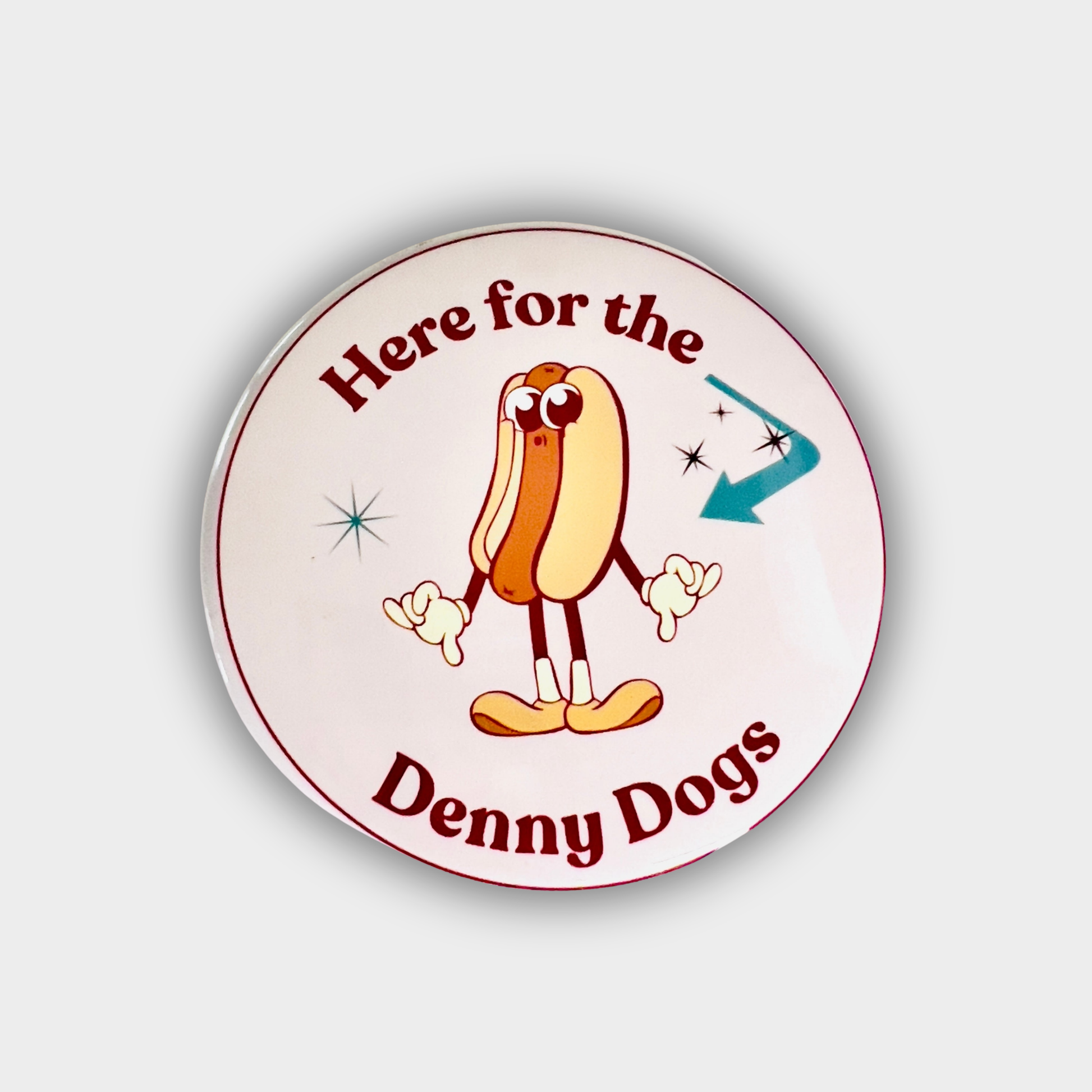Round button with cartoon hot dog and text 'Here for the Denny Dogs' on a light gray background