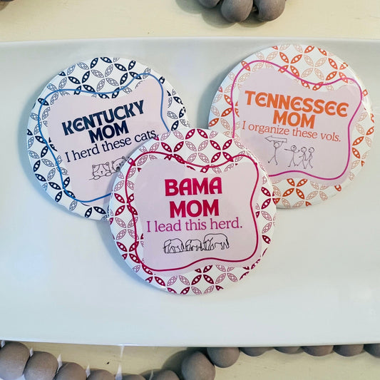 Three college buttons with humorous text about different college moms on a white surface with gray beads.