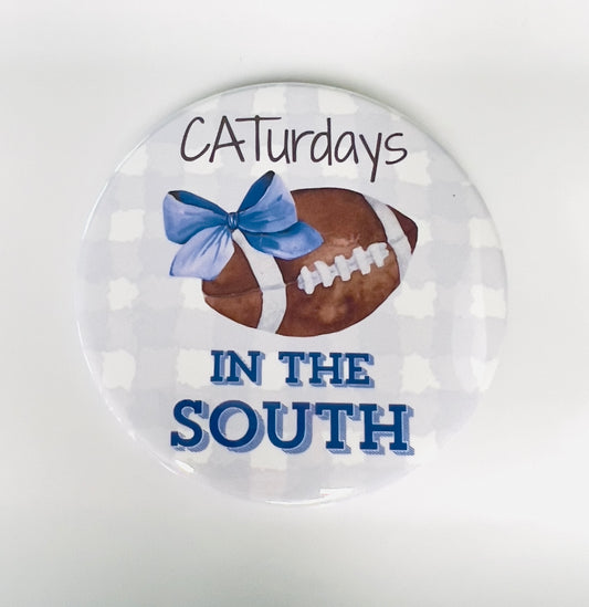 Round button with football, blue bow, and text 'Caturdays IN THE SOUTH' on a light blue and white checked background