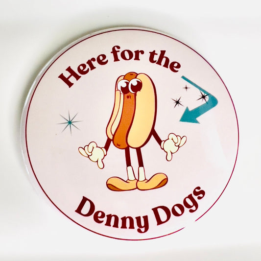 Circular sign with cartoon hot dog character and text 'Here for the Denny Dogs' on a white background