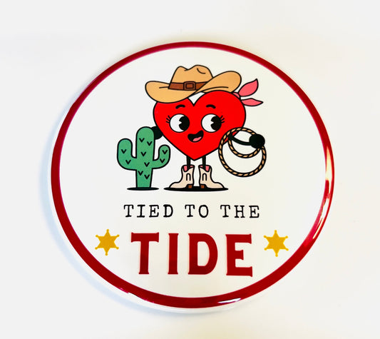 Round button with a cartoon heart character, cactus, cowboy hat and text 'Tied to the Tide' on a white background.