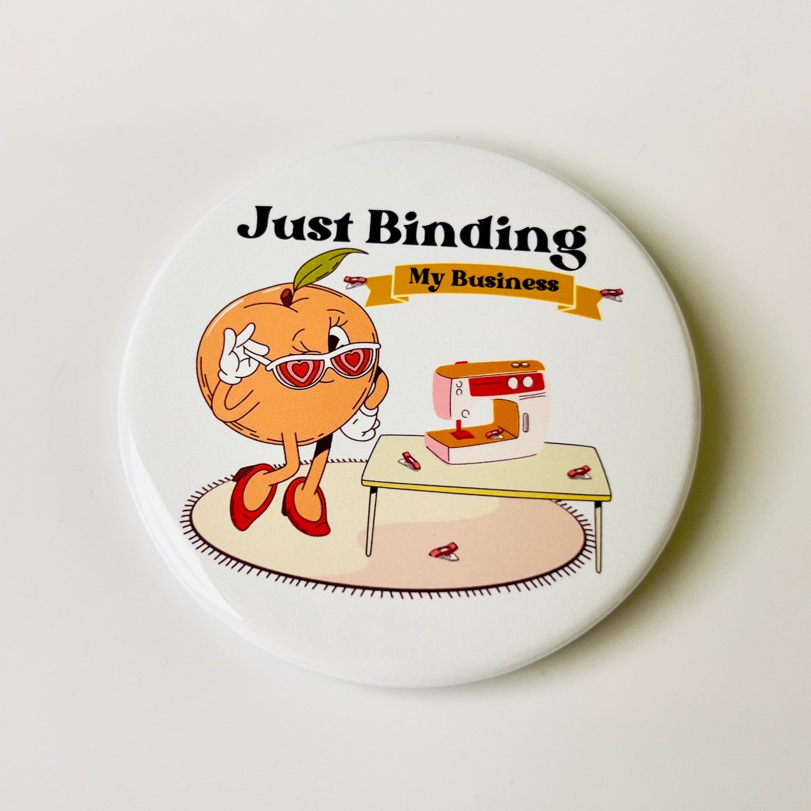 Button badge with cartoon peach character and sewing machine, text 'Just Binding My Business' on a light background.