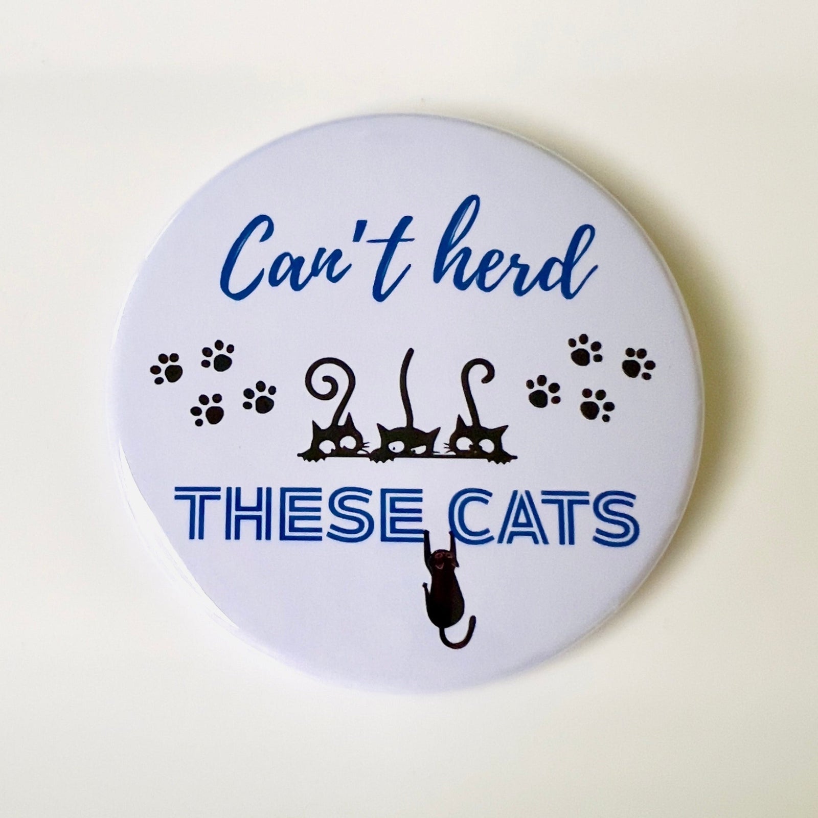 Button badge with text 'Can't herd these cats' and cat illustrations on a white background