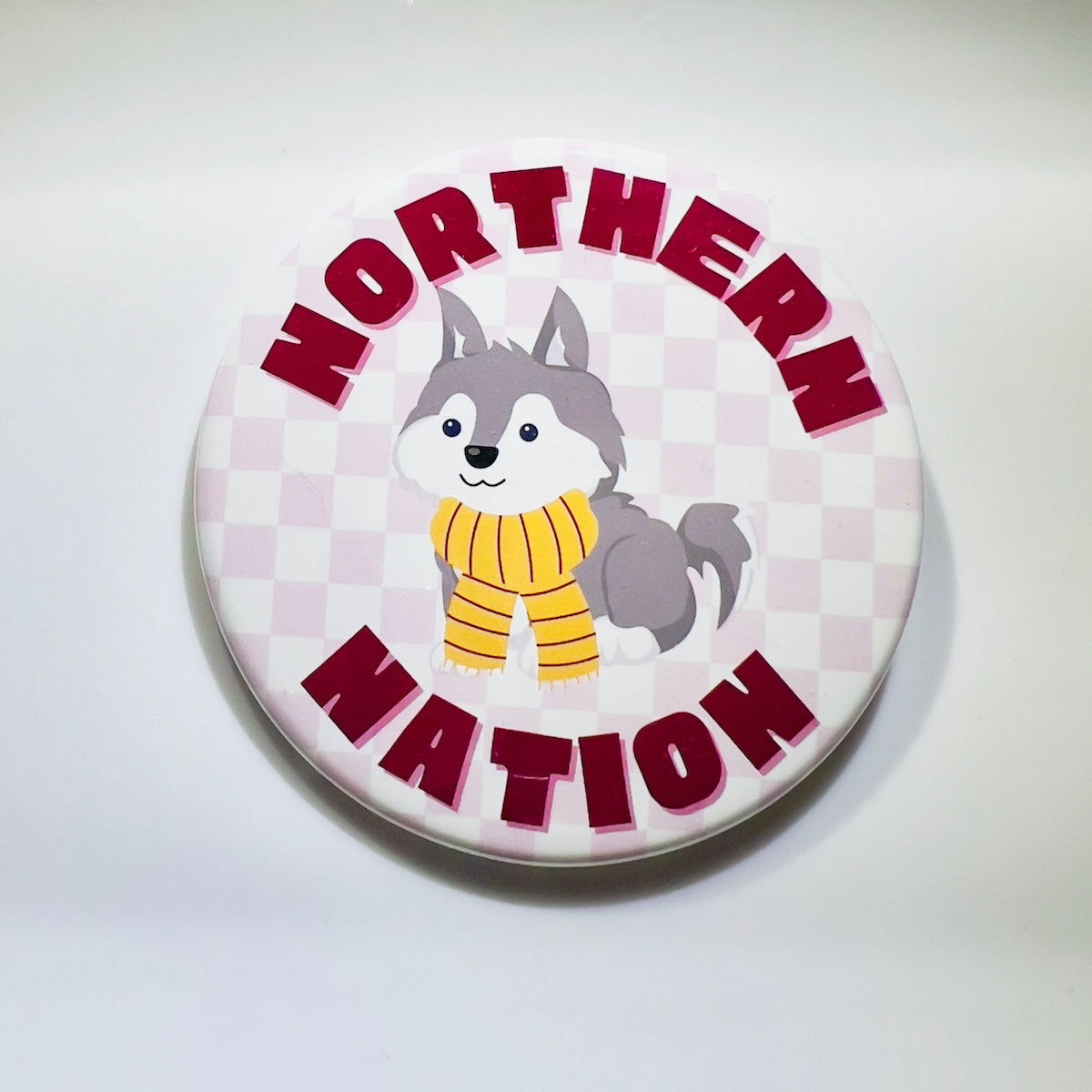 Circular button with wolf pup wearing yellow scarf on pink and white checkered background