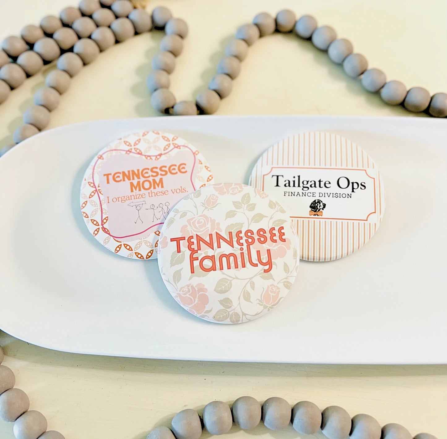 Three circular Tennessee buttons with text on a white tray, surrounded by wooden beads.