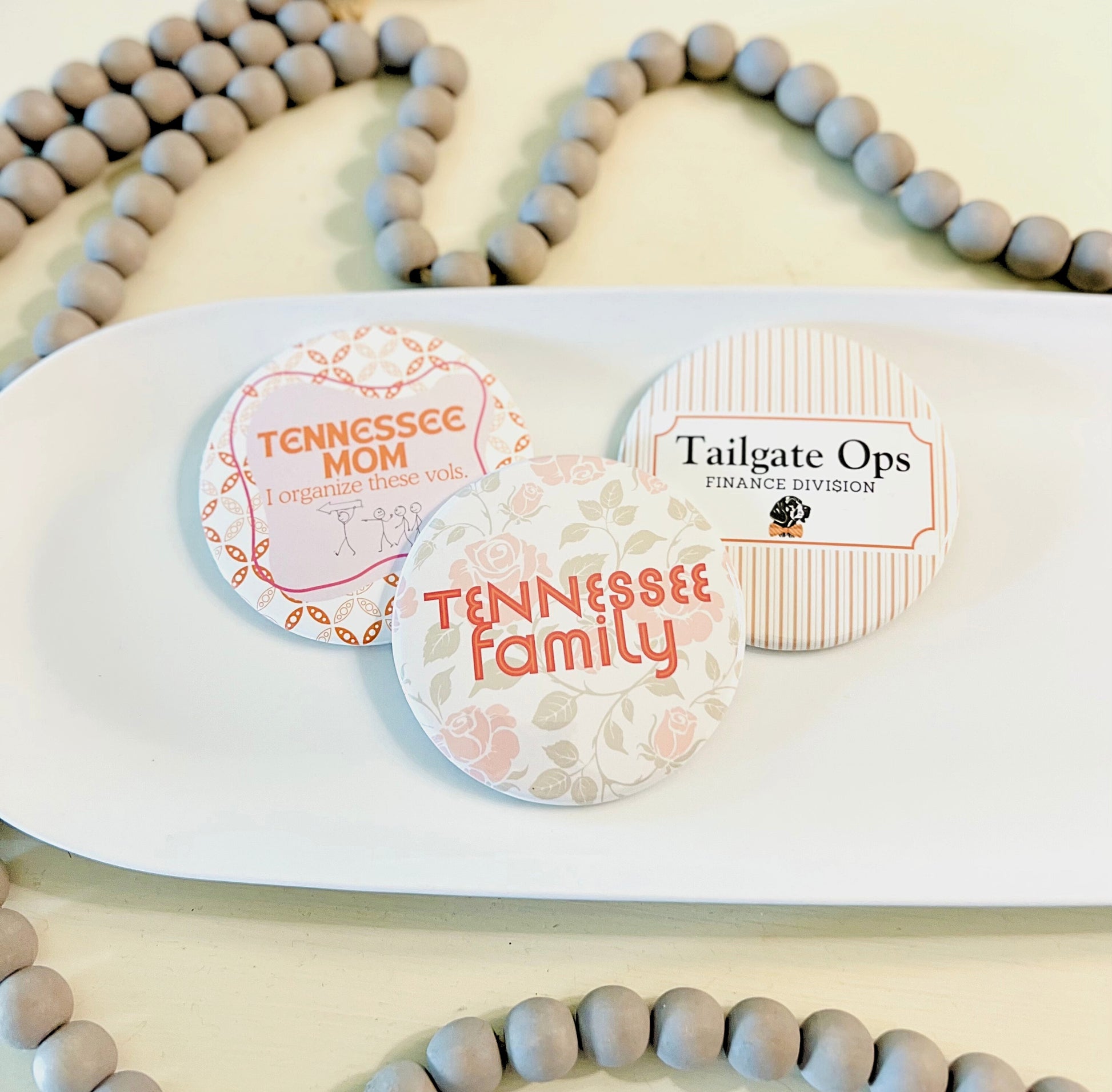 Three circular Tennessee buttons with text on a white tray, surrounded by wooden beads.