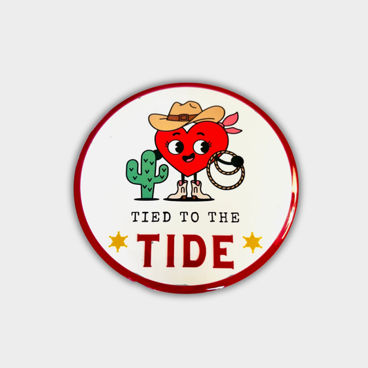 Round button with a cartoon heart character, cactus, cowboy hat and text 'Tied to the Tide' on a white background.